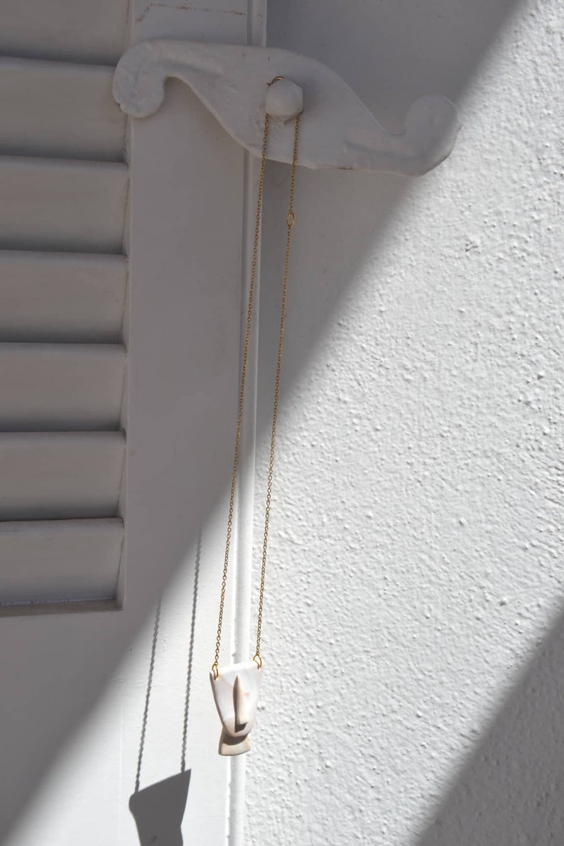 Op de afbeelding: Een gouden ketting met een witte keramische hanger in de vorm van een gezicht. De hanger heeft een eenvoudig ontwerp met een kleine neus en mond.