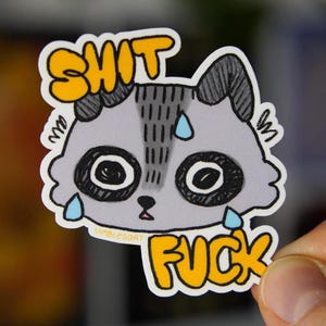 May include: A die-cut sticker featuring a cartoon raccoon with a distressed expression. The raccoon is grey and white with large black eyes and yellow text that reads "SHIT FUCK". The sticker has a white border.