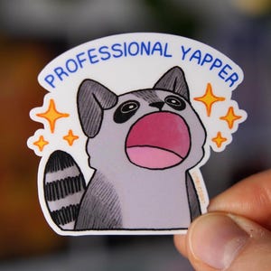Puede incluir: Una pegatina con un mapache de dibujos animados con la boca abierta, rodeado de estrellas. El texto "PROFESSIONAL YAPPER" está sobre el mapache. El mapache es gris, blanco y negro con la boca rosa.