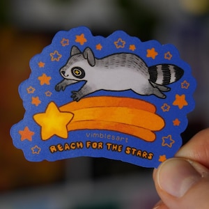 Puede incluir: Una pegatina azul con un mapache de dibujos animados saltando sobre un cometa naranja y una estrella amarilla. La pegatina tiene estrellas naranjas en el borde y el texto "Reach for the Stars".