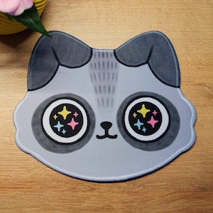 May include: A grey cat-shaped mouse mat with large eyes featuring colourful starburst designs. The mouse mat has a light blue border and is on a wooden surface. A pink keyboard and a yellow vase with a pink flower are in the background.