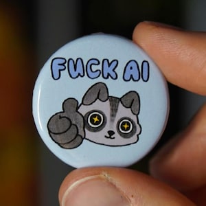 Könnte beinhalten: Ein hellblauer Knopf mit dem Text "FUCK AI" in blauen Buchstaben. Der Knopf zeigt einen Cartoon-Waschbären, der einen Daumen hoch zeigt. Der Waschbär hat graues Fell und große Augen.
