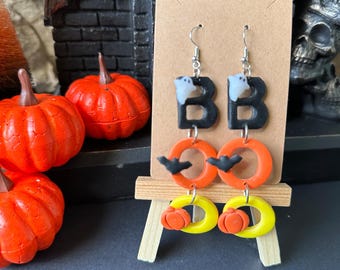May include: A pair of Halloween-themed earrings. The first pair features black letter Bs with white ghosts inside. The second pair features orange circles with black bats and yellow circles with orange pumpkins.
