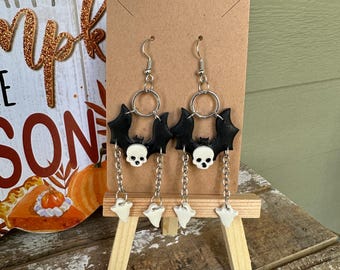 Puede incluir: Un par de pendientes de murciélago negro con dijes de calavera blanca y dijes de fantasma blanco que cuelgan de cadenas plateadas.