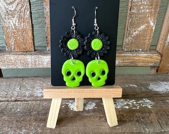 Puede incluir: Un par de pendientes colgantes de calavera verde y flor negra. Los pendientes están en una tarjeta de exhibición negra.