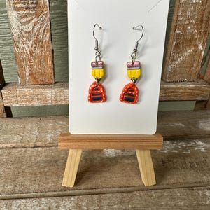Puede incluir: Un par de pendientes hechos a mano con temática escolar. Cada pendiente tiene una parte superior de lápiz amarillo con una goma de borrar rosa y un dije de mochila rojo y naranja. Los pendientes se exhiben en una tarjeta blanca.