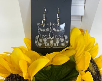 May include: A pair of clear acrylic cat earrings with a floral design. The earrings are hanging from a black card.