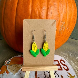 May include: A pair of dangle earrings featuring a yellow corn cob with green husks. The earrings are displayed on a brown card stand.