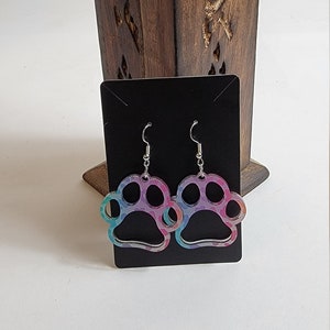 Peut inclure: Une paire de boucles d'oreilles pendantes avec un motif de patte d'animal. Les pattes sont en acrylique iridescent et présentent un motif multicolore. Les boucles d'oreilles sont argentées et ont une fermeture à crochet.