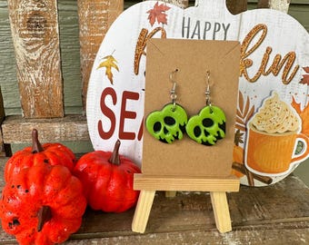 Puede incluir: Pendientes verdes en forma de calavera con detalles negros que cuelgan de ganchos plateados. Los pendientes se muestran en un pequeño caballete de madera frente a un cartel de calabaza blanca que dice "Happy Fall".