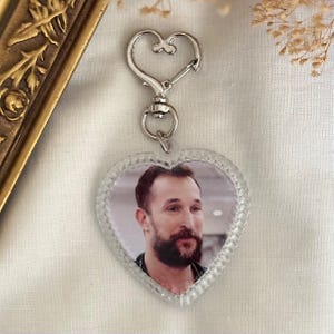 Puede incluir: Llavero acr&iacute;lico transparente en forma de coraz&oacute;n con cierre plateado. El llavero presenta una fotograf&iacute;a de un hombre con barba. El colgante en forma de coraz&oacute;n tiene un borde texturizado.