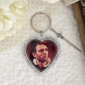 May include: A heart-shaped keychain featuring a portrait of a person with dark hair and a white ruffled collar. The keychain includes a silver-toned metal ring and a small, detailed silver-toned sword charm attached to the side of the ring.
