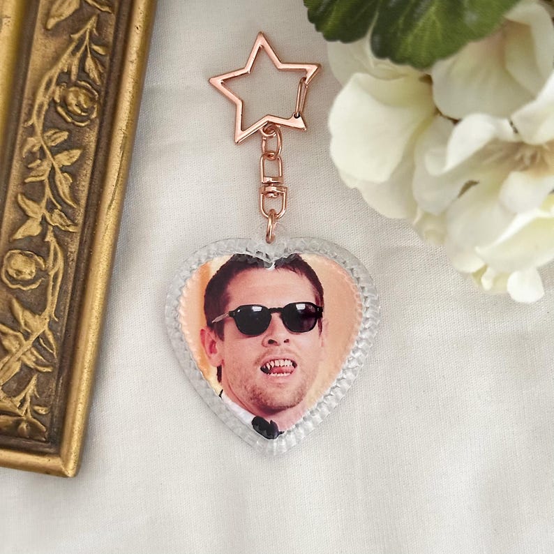 May include: A heart-shaped keychain featuring a man wearing sunglasses. The keychain has a clear, textured border and a rose gold star-shaped clasp. The background includes a gold-colored frame and white flowers.