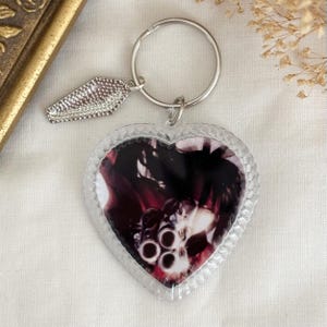 May include: A clear heart-shaped keyring with a black and white photo of a gun inside. The keyring has a silver metal coffin charm attached to a silver metal ring.