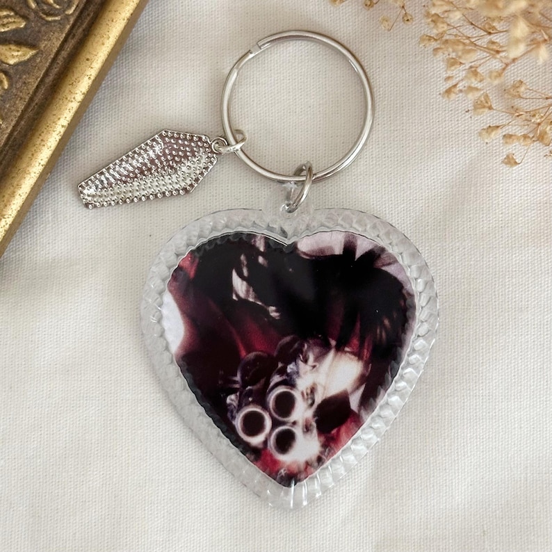 May include: A clear heart-shaped keyring with a black and white photo of a gun inside. The keyring has a silver metal coffin charm attached to a silver metal ring.