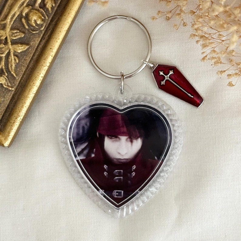 May include: A clear heart-shaped keyring with a photo of a character with red hair and a red and black outfit. The keyring also has a silver key ring with a red coffin charm with a silver cross.