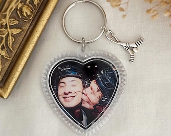 Könnte beinhalten: Herzförmiger Schlüsselanhänger mit einem Foto von zwei Personen mit Hockeyhelmen. Der Schlüsselanhänger hat einen silbernen Ring und einen Hockey-Schläger-Anhänger. Der herzförmige Anhänger hat einen klaren, strukturierten Rand.