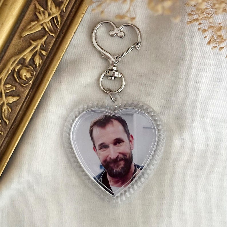 Puede incluir: Un llavero transparente en forma de coraz&oacute;n con un cierre plateado. El llavero presenta una foto de un hombre con barba. El marco en forma de coraz&oacute;n tiene un borde texturizado y con cuentas. El fondo es una tela blanquecina.