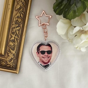 May include: A clear heart-shaped keychain with a photo of a smiling person wearing sunglasses. The keychain has a rose gold star-shaped top and a metal clasp. The background is a white surface with flowers and a gold frame.