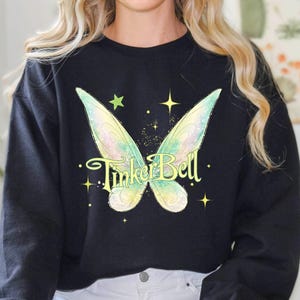 May include: Black sweatshirt featuring a whimsical design of fairy wings and the text "Tinker Bell" in a playful font. The wings are rendered in shades of green and yellow, with sparkling stars. A comfortable and stylish top.