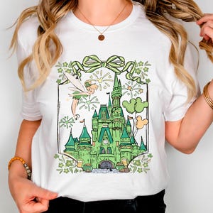 May include: White t-shirt featuring a green castle illustration with a fairy, fireworks, and balloons. The design includes a green bow and floral accents. The castle has multiple towers and a detailed facade.