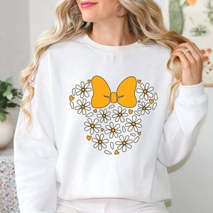 Puede incluir: Sudadera blanca con un lazo amarillo y margaritas blancas que forman la forma de la cabeza de Mickey Mouse.