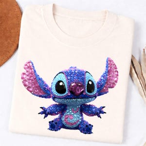 K&ouml;nnte beinhalten: Cremefarbenes T-Shirt mit einem funkelnden, mit Juwelen besetzten Stitch-Charakter. Die Figur ist haupts&auml;chlich blau mit rosa Akzenten an Ohren und Brust. Gro&szlig;e schwarze Augen und ein rosa Edelstein auf der Nase vervollst&auml;ndigen das Design. Das T-Shirt ist gefaltet.