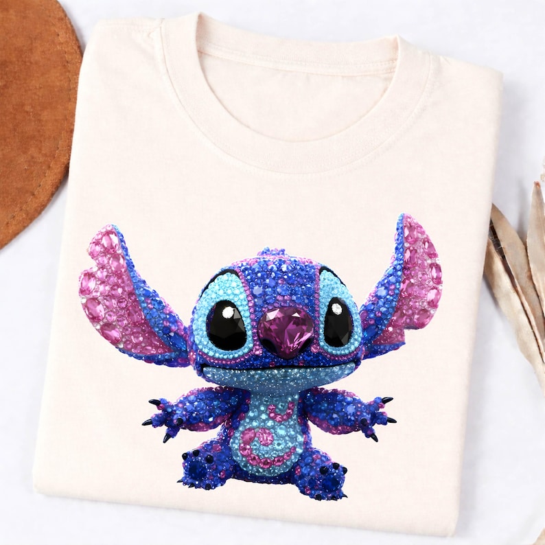 K&ouml;nnte beinhalten: Cremefarbenes T-Shirt mit einem funkelnden, mit Juwelen besetzten Stitch-Charakter. Die Figur ist haupts&auml;chlich blau mit rosa Akzenten an Ohren und Brust. Gro&szlig;e schwarze Augen und ein rosa Edelstein auf der Nase vervollst&auml;ndigen das Design. Das T-Shirt ist gefaltet.