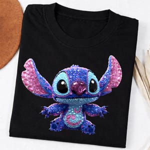 K&ouml;nnte beinhalten: Schwarzes T-Shirt mit Rundhalsausschnitt und einem glitzernden Cartoon-Charakter in Blau und Pink. Die Figur hat gro&szlig;e Augen, eine lila Nase und gro&szlig;e Ohren. Das Design ist auf dem T-Shirt zentriert.
