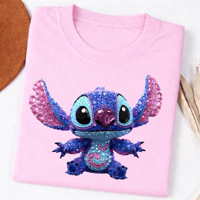 K&ouml;nnte beinhalten: Ein hellrosa T-Shirt mit einer glitzernden, mit Juwelen besetzten Cartoonfigur in Blau- und Rosat&ouml;nen. Die Figur hat gro&szlig;e schwarze Augen und einen rosa Edelstein auf der Nase. Das T-Shirt ist auf einer wei&szlig;en Oberfl&auml;che gefaltet.