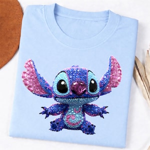 K&ouml;nnte beinhalten: Hellblaues T-Shirt mit einem funkelnden Stitch-Charakterdesign. Die Figur ist haupts&auml;chlich blau mit rosa Akzenten an Ohren und Brust. Das Shirt besteht aus einem weichen Material und ist f&uuml;r den Freizeitgebrauch geeignet.