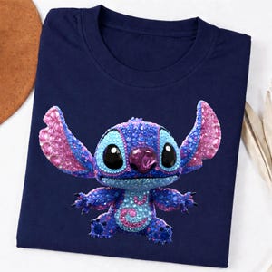 K&ouml;nnte beinhalten: Marineblaues T-Shirt mit einem funkelnden Stitch-Cartoon-Charakter. Die Figur ist in Blau-, Lila- und Rosat&ouml;nen gehalten, mit gro&szlig;en schwarzen Augen und einer juwelenartigen Nase. Das Shirt hat einen klassischen Rundhalsausschnitt.