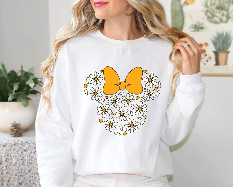 Puede incluir: Sudadera blanca con un lazo amarillo y margaritas blancas que forman la forma de la cabeza de Mickey Mouse.