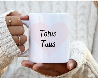 Könnte beinhalten: Weiße Keramik-Tasse mit einem hellrosa Herz und den Worten "Totus Tuus" in schwarzer Schrift. Die Tasse wird von einer Person gehalten, die einen weißen Pullover trägt. Ein schlichtes und elegantes Design für den täglichen Gebrauch.