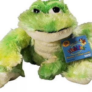 May include: A plush frog toy in shades of green and yellow with a cream-colored belly. The frog has black eyes and a tag that reads "Webkinz" and "Adopt a pet, discover a virtual world!" from GANZ.