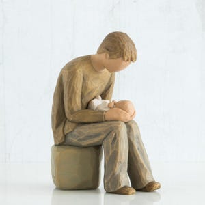 May include: A brown figurine of a person seated on a small square stool, cradling a baby. The person is wearing a long-sleeved shirt and pants. The baby is wrapped in a white blanket. The figurine has a carved wood-like texture.
