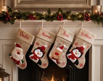 May include: Four beige fabric Christmas stockings hanging in a row. Each stocking features a personalized name embroidered in red script: Dada, Mama, Aria, and Willow. The stockings are decorated with embroidered holiday characters: Santa, a penguin, a reindeer, and another penguin, all accented with red snowflakes.