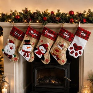 May include: Five Christmas stockings hanging in a row, each featuring a red and black plaid cuff with embroidered names: Dad, Mum, Gracie, Lily, and Jax. The stockings display festive appliqués of Santa, a snowman, a penguin, a reindeer, and a paw print.