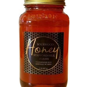 May include: A glass jar of honey with a gold lid and a black label. The label says "Sourwood Honey Stafford Hill Farms 115 Edgewood Rd S Asheville, NC 28803 2 LB (907 g)"
