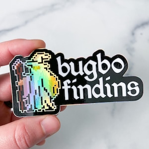 May include: A black and white sticker with a holographic finish. The sticker features a pixelated image of a bugbear holding a staff and the text "bugbo findins".