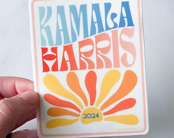 May include: A sticker with a retro-style design featuring the text "KAMALA HARRIS 2024" in a colorful, wavy font. The design includes a stylized sunburst with orange, yellow, and red rays.