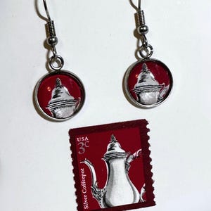May include: A pair of silver-toned dangle earrings featuring circular charms with a red background and a white illustration of a silver coffeepot. Below the earrings is a rectangular postage stamp with the text USA 3c Silver Coffeepot.