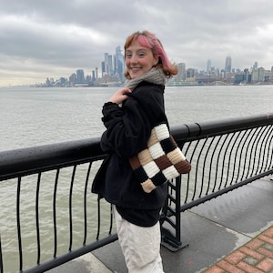 May include: A person with pink hair is standing in front of a city skyline. They are wearing a black jacket, a white scarf, and white pants. They are holding a brown, black, and white checkered crocheted bag.
