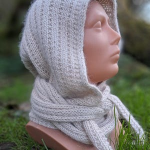 May include: A light beige knitted hood and scarf set. The hood is attached to a scarf that wraps around the neck. The set is made of a soft, textured yarn. The background is blurred, with green grass and foliage.
