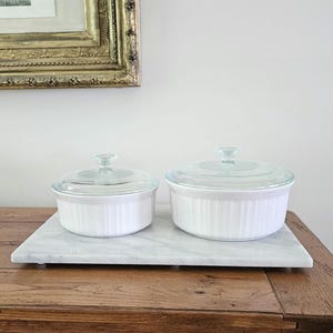 May include: Two round white ceramic baking dishes with vertical ribbed sides and clear glass lids. The dishes are of different sizes, both featuring a smooth finish and a knob handle on the center of each lid, providing a clean and functional design.