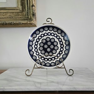 May include: A round ceramic plate featuring a repeating pattern of dark blue and white concentric circles, displayed on a decorative bronze-toned metal stand. The design consists of small circles arranged in circular rows across the surface.