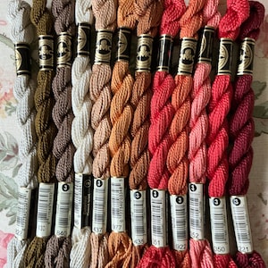May include: A collection of DMC embroidery floss in various shades of brown, peach, and red. The floss is neatly arranged, with each skein labeled with a number. The colors range from light beige to deep burgundy, perfect for embroidery projects.