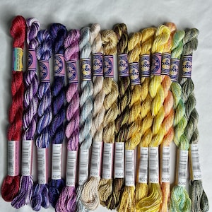 May include: A collection of thirteen skeins of DMC Cotton Perle size 5 embroidery thread in a gradient of colors including red, purple, blue, pink, light blue, beige, gold, yellow, orange, and green, each with a paper label and barcode.