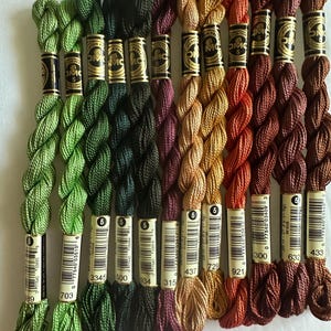 May include: A collection of ten skeins of DMC Coton Perlé size 5 embroidery thread in a gradient of colors including light green, dark green, deep purple, gold, orange, and various shades of brown, each with a paper label showing the color number.