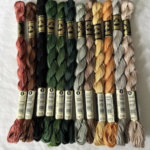 May include: A collection of twelve DMC Cotton Perle size 5 embroidery threads arranged in a row. The skeins feature a variety of colors including rust, brown, black, dark green, forest green, sage, tan, gold, yellow, silver, and beige.
